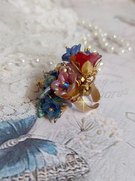 Bague Bel Air mont&eacute;e avec des fleurs lucites peintes &agrave; la main aux couleurs : Bleu, Rose, Rouge, Argent, Blanc avec feuilles ondul&eacute;es Vert Cuivr&eacute; &agrave; laquelle s&rsquo;ajoute perles Dor&eacute;es en Verre