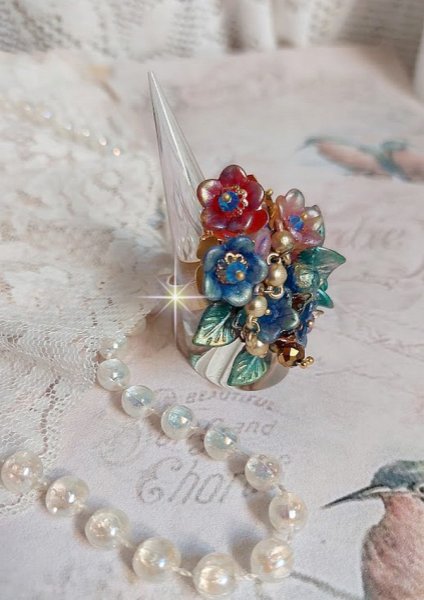 Bague Bel Air mont&eacute;e avec des fleurs lucites peintes &agrave; la main aux couleurs : Bleu, Rose, Rouge, Argent, Blanc avec feuilles ondul&eacute;es Vert Cuivr&eacute; &agrave; laquelle s&rsquo;ajoute perles Dor&eacute;es en Verre