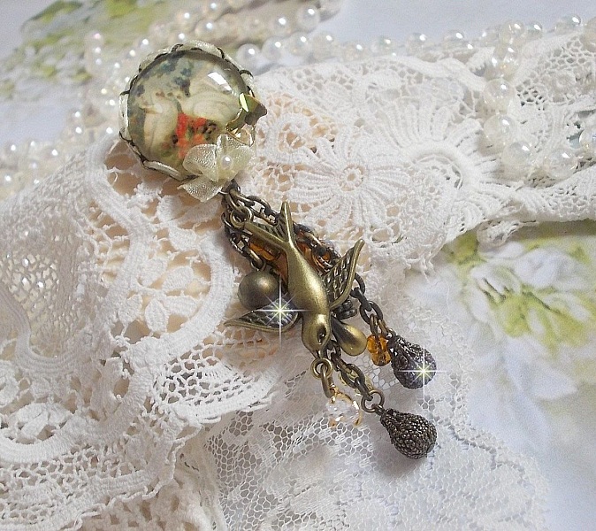 Broche Colombes aux plumages blancs avec des breloques filigran&eacute;es gouttes et lisses, perles rondes et n&oelig;ud en cristal, ruban organza cr&egrave;me, pendentif plaqu&eacute; or avec un zircon sur une broche en m&eacute;tal