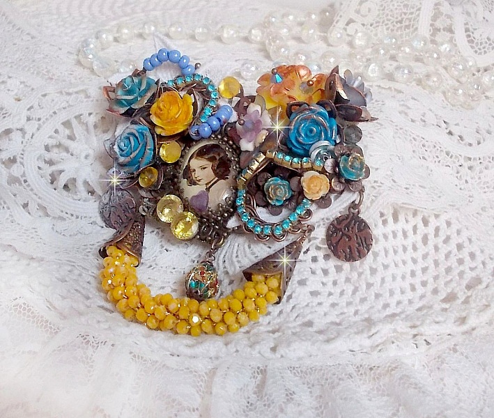 Broche Eclats P&eacute;tillants d&rsquo;inspiration Baroque, orn&eacute;e de fleurs en r&eacute;sine autour d&rsquo;un cabochon strass&eacute;s et perl&eacute;s sur estampes, cristaux, breloques, perle cloisonn&eacute;e, perles jaunes en verre et roses. 