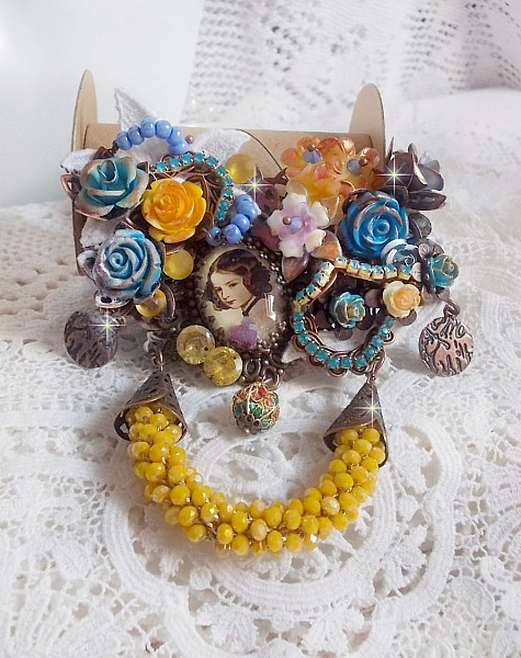 Broche Eclats P&eacute;tillants d&rsquo;inspiration Baroque, orn&eacute;e de fleurs en r&eacute;sine autour d&rsquo;un cabochon strass&eacute;s et perl&eacute;s sur estampes, cristaux, breloques, perle cloisonn&eacute;e, perles jaunes en verre et roses. 